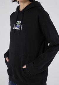 Černá mikina s kapucí a přední kapsou, s grafickým designem a textem "THE JOKER." Vyrobena z měkké látky, se stahovací šňůrkou a žebrovanými manžetami.