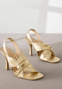 Gouden slingback hakken met een gedraaide stofontwerp, open teen en hoge stiletto hak. Glad, glanzend textuur met een nude binnenzool.