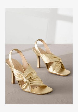 Zlatne potpetice s remenčićem, s uvijenim dizajnom tkanine, otvorenim prstima i visokim stiklom. Glatka, sjajna tekstura s nude uloškom.