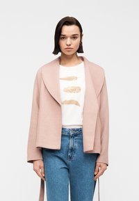 Γυναίκα με κοντά σκουρόχρωμα μαλλιά φοράει ένα απαλό ροζ σακάκι πάνω από ένα λευκό T-shirt με εκτυπώσεις φτερών και μπλε τζιν ψηλόμεσα, στέκεται μπροστά σε λευκό φόντο.