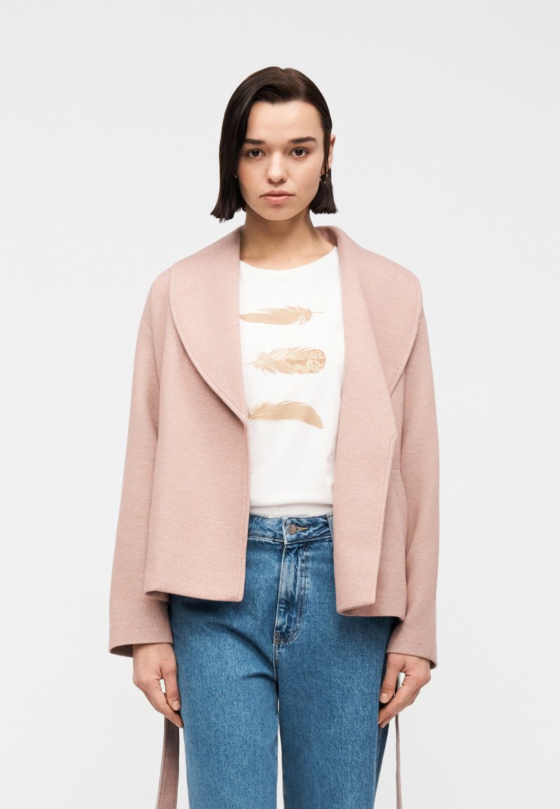 Γυναίκα με κοντά σκουρόχρωμα μαλλιά φοράει ένα απαλό ροζ σακάκι πάνω από ένα λευκό T-shirt με εκτυπώσεις φτερών και μπλε τζιν ψηλόμεσα, στέκεται μπροστά σε λευκό φόντο.