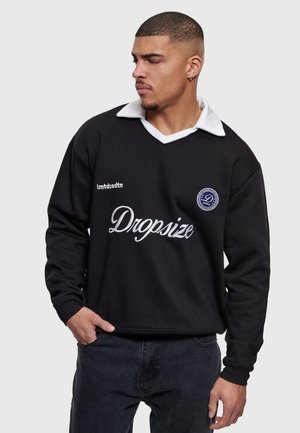 Mann trägt schwarzen Pullover mit weißem Kragen und dem Schriftzug "Dropsize", schaut seitlich und hat eine Hand in der Tasche vor einfachem Hintergrund.