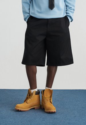 Čovjek u crnim kratkim hlačama do koljena, bijelim čarapama i sivim radnim čizmama stoji na plavom tepihu.