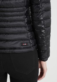 Nahaufnahme einer Person, die eine schwarze gesteppte Jacke und schwarze Hosen trägt, wobei die Rückseite und die Armdetails der Jacke gezeigt werden.