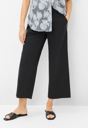 Personne portant un pantalon noir large court, des sandales noires à enfiler et une chemise blanche transparente à motif floral avec des rayures verticales.