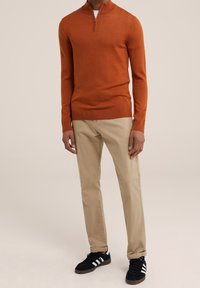 Orange zip-up tröja, beige chinos, svarta sneakers med vita ränder. Tröjan har ribbade ärmslut och nederkant, slät textur.
