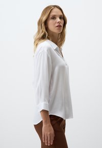 Chemisier blanc à manches longues avec col, en tissu texturé et poignets retroussés, associé à un pantalon marron sur un fond neutre.