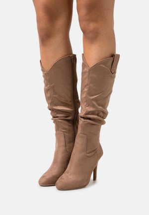 Jambes portant des bottes à talons hauts en daim beige mi-mollet, avec une fermeture éclair latérale et un shaft légèrement froissé, sur un fond uni.