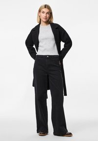 Abrigo largo negro sobre un suéter de canalé gris claro, combinado con pantalones de pierna ancha negros y botas gruesas de tacón marrón oscuro. Diseño simple y moderno.