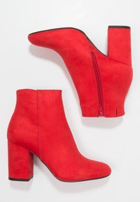 Czerwone zamszowe botki za kostkę z ostro zakończonym noskiem, tylnym zamkiem i słupkowym obcasem. Projekt charakteryzuje się gładką teksturą i prostymi liniami.