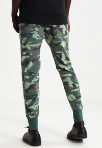 Kamouflage sweatingbyxor i gröna och svarta nyanser, tapered ben design, elastiska muddar och en mjuk tygstruktur. Svarta skor synliga nedanför.