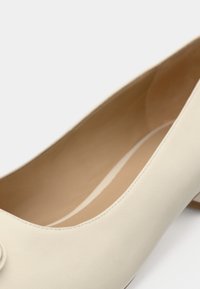 Ballerine en cuir beige avec un bout arrondi, des détails de couture et une texture lisse, ornée d'un nœud décoratif à l'avant.