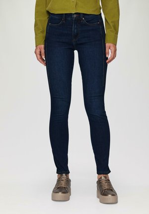 Jeans skinny bleu foncé en denim, avec une taille haute, des détails de couture subtils, et associés à des baskets métalliques.