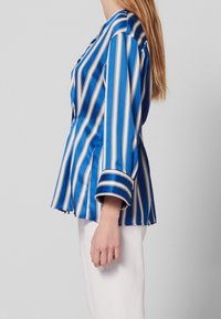 Modrá a bílá pruhovaná saténová blůza s volným střihem, širokými rukávy a bočními rozparky, kombinovaná s bílými kalhotami. Hladká textura.