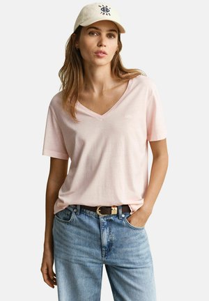 Vrouw die een lichtroze T-shirt met V-hals draagt, lichtblauwe spijkerbroek met een bruine riem en een beige baseballpet, staand met één hand in de zak.