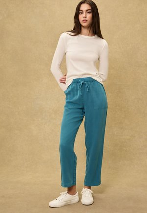 Ung kvinde står med hænderne bag ryggen iført cremefarvet top med lange ærmer, teal bukser med snørre og hvide sneakers på beige baggrund.