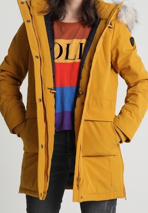Parka jaune avec capuche bordée de fourrure, fermeture éclair et plusieurs poches avant, portée sur une chemise rayée multicolore.