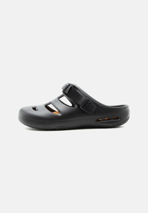 Juodos spalvos lengvai užsidedančios basutės su reguliuojamu dirželiu, atvirais šonais ir uždaru priekiniu galiuku, skirtos laisvalaikiui ir lauko veiklai.