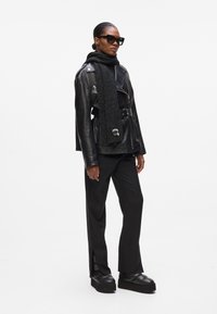 Schwarze Lederbikerjacke mit silberner Hardware, schwarzes Rollkragenoberteil, strukturierter schwarzer Schal, schwarze weit geschnittene Hose mit seitlichen Schlitzen und Plateauschuhe.