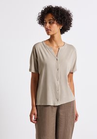 Beige Kurzarmhemd mit rundem Ausschnitt und Knopfleiste. Leichter Stoff, glatte Textur. Kombiniert mit lockeren braunen Leinenhosen.