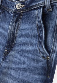 Pantalones vaqueros de mezclilla en azul oscuro, con un diseño clásico de cinco bolsillos, herrajes de latón y detalles de costura visibles en la cintura y los bolsillos.