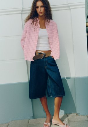 Jeune femme aux cheveux bouclés portant une chemise rose à carreaux, un débardeur blanc, un short en jean large, une ceinture marron et des mules à talons roses, debout contre un mur blanc.