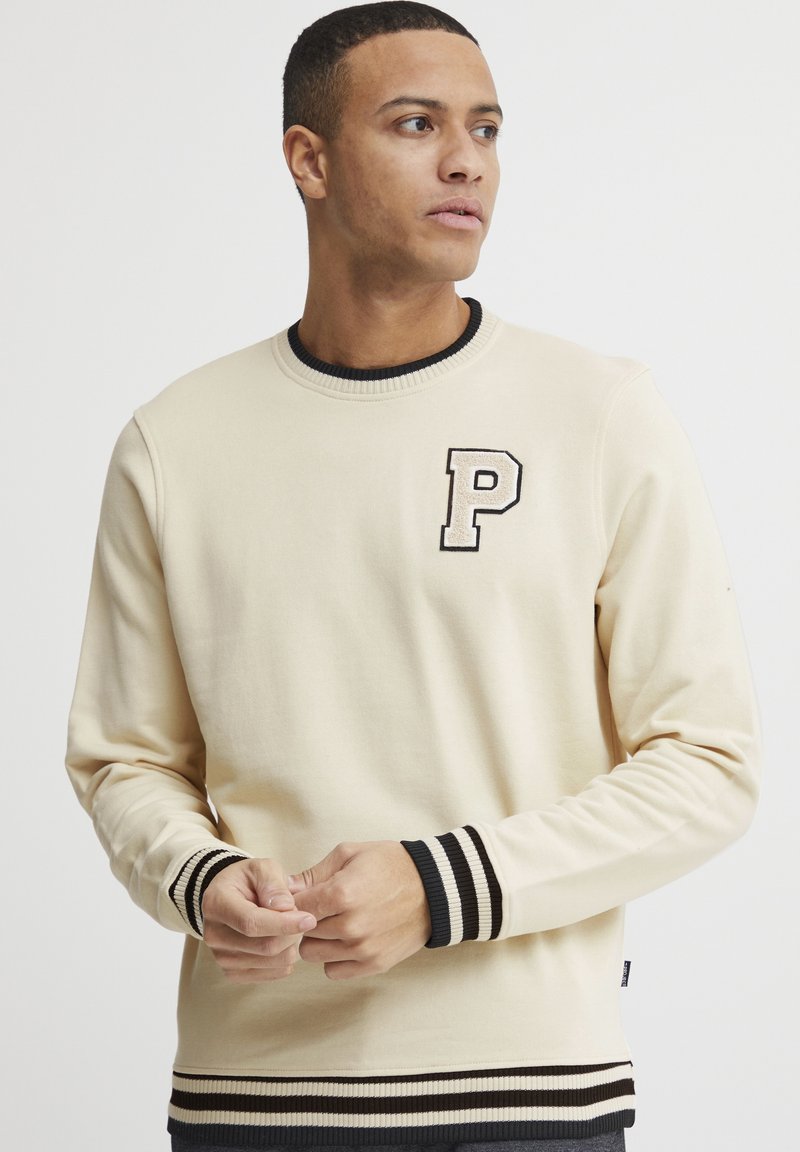 Sudadera beige de algodón, con un emblema negro en forma de 'P', puños y bajo acanalados con rayas en blanco y negro, y cuello redondo.