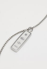Pendentif en argent avec une chaîne, comportant des logos et des symboles gravés, incluant une balle de tennis et le mot "PARIS". Texture lisse et métallique.