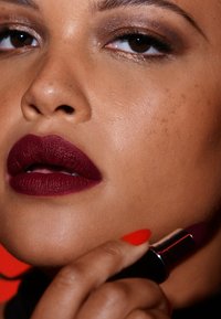 Donkerburgundy lippenstift aangebracht op volle lippen. De lippen vertonen een matte textuur. Een hand met felrode nagels houdt een lippenstiftbuisje vast.