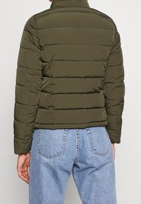 Olivengrøn puffjakke med høj krave, quiltet tekstur og kort længde. Kombineret med lyseblå denimjeans.