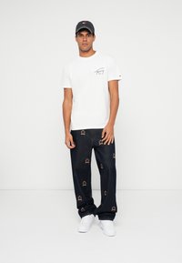 Λευκό T-shirt με λογότυπο στο στήθος, συνδυασμένο με σκούρο τζιν παντελόνι με κεντημένα σχέδια στέμματος. Λευκά αθλητικά παπούτσια και γκρι καπέλο.