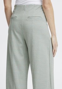 Pantalons à carreaux verts et marron avec une taille haute, des poches latérales et des plis marqués, en tissu lisse et texturé.