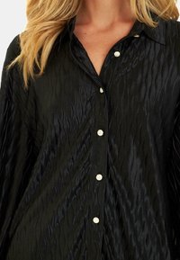 Blusa negra hecha de una tela texturizada con un patrón ondulado. Presenta un cuello clásico y botones en estilo perla con un brillo sutil.
