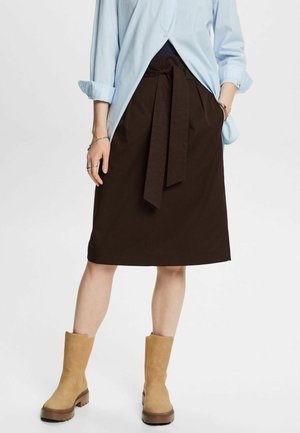 Femme portant une jupe marron foncé jusqu'aux genoux avec une ceinture nouée à la taille, une chemise bleu clair aux manches retroussées, et des bottines couleur beige.