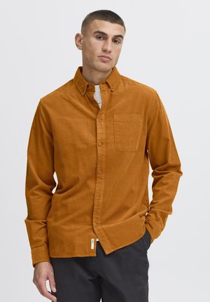 Ung mand iført en sennepsfarvet ribfløjls-skjorte over en hvid T-shirt, med sorte bukser, stående mod en ensfarvet lys baggrund.