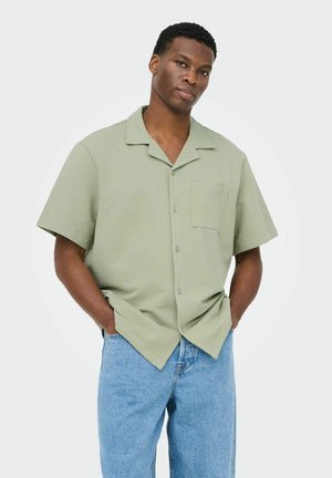 Uomo che indossa una camicia a maniche corte verde chiaro con taschino sul petto e jeans azzurri, in piedi con le mani nelle tasche contro uno sfondo semplice.