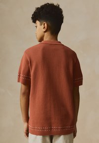 Maglione a maniche corte color ruggine con motivo a texture, dotato di colletto e dettagli di cuciture bianche. Vista posteriore.