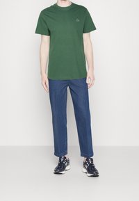 Man som bär en mörkgrön t-shirt med korta ärmar, blå raka jeans och svartvita sneakers, stående mot en enkel vit bakgrund.