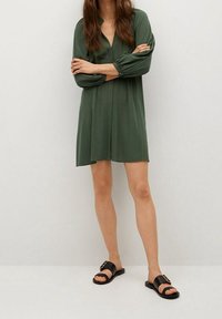 Vihreä lyhyt mekko, jossa on V-pääntie, pitkät hihat ja pleatattu muotoilu. Yhdistetty mustiin tasapohjaisiin sandaaleihin, joissa on kultainen solki.