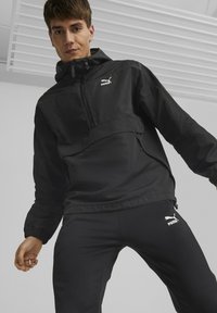 Svart Puma vindjakke med halv glidelås, justerbar hette og elastiske mansjetter, sammen med svarte tettsittende bukser med logo på låret.