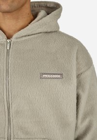 Jasnoszara bluza polarowa z kapturem zapinana na zamek z przodu, z naszywką z logo oraz sznurkiem w kapturze, prezentująca miękką, teksturowaną powierzchnię.