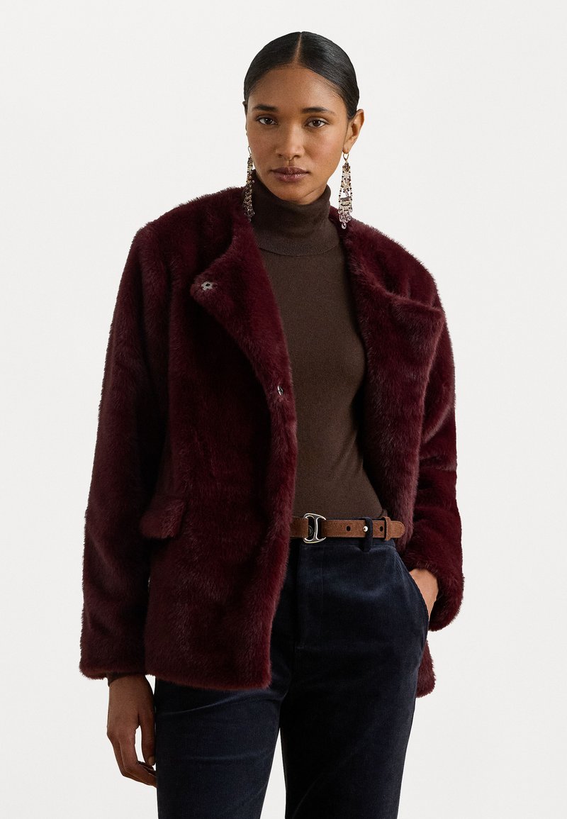 Burgundi színű műszőrme dzseki laza szabással, kihajtott gallérral, két elülső zsebbel és puha, texturált felülettel. Sötét garbó fölé viselve.