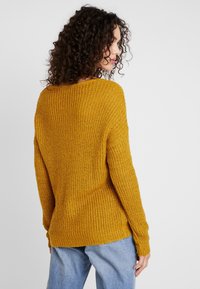 Hořčicově žlutý pletený svetr s volným střihem, žebrovanou strukturou a širokým výstřihem, spárovaný se světle modrými džíny.