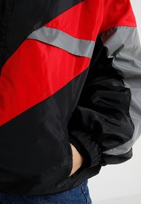 Giacca a vento rossa e nera con blocchi di colore diagonali, strisce riflettenti grigie, polsini elasticizzati e vestibilità rilassata.