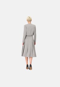 Robe grise avec un corsage ajusté et une jupe plissée, manches longues et col montant. Portée avec des chaussures à talons hauts noires.