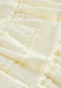 Getextureerde crèmekleurige stof met gelaagde ruches en kantafwerking, die een zachte, geweven uitstraling en delicate details door het hele ontwerp toont.