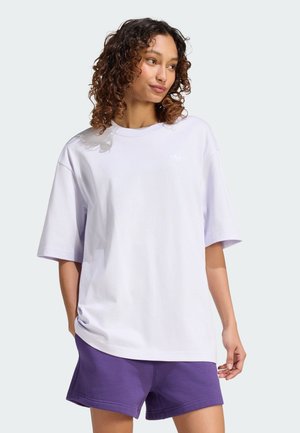 Kvinde med krøllet hår iført en løs hvid T-shirt og lilla shorts, stående mod en ensfarvet lys baggrund, kigger til siden.