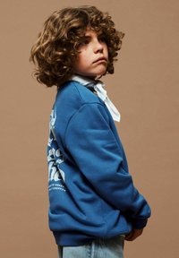 Enfant aux cheveux bouclés portant un sweat-shirt bleu avec un texte blanc au dos, un foulard blanc autour du cou et un jean bleu clair, sur un fond beige.