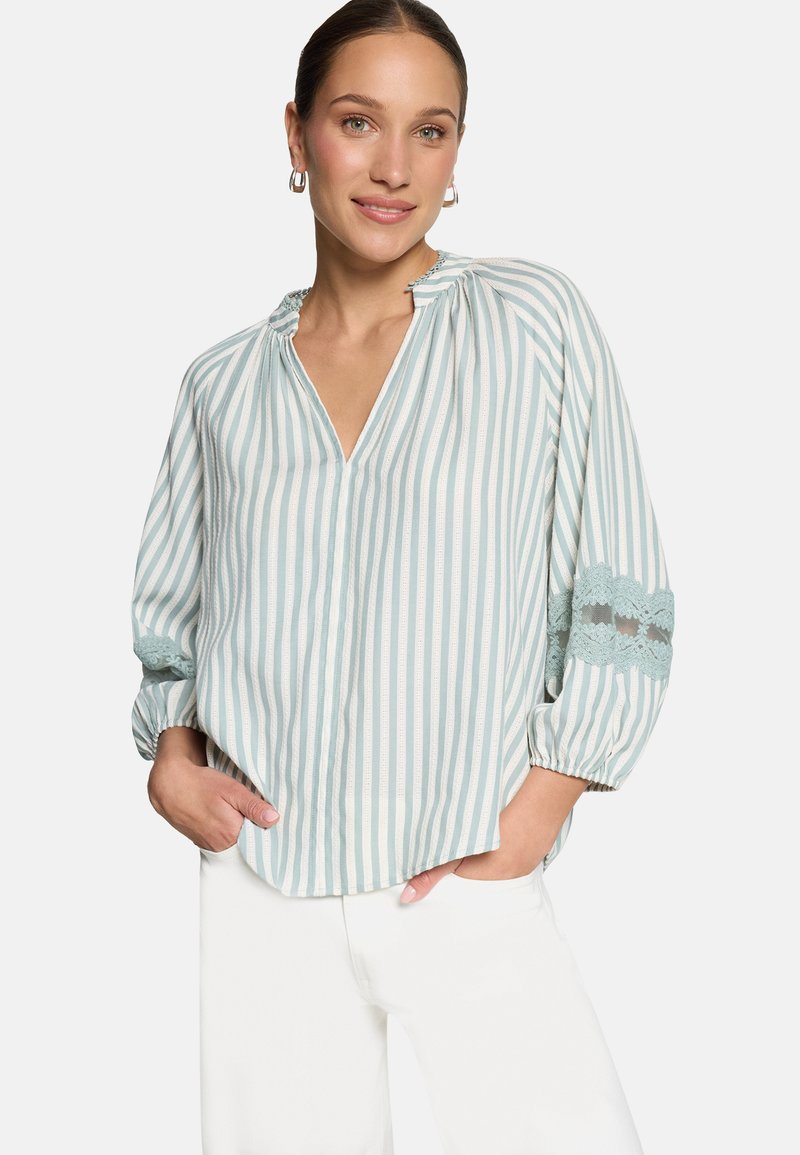 Žena oblečená v svetlozeleno-bielem blúzke s vertikálnymi pruhmi a čipkovým detailom na rukávoch, v bielych nohaviciach, usmievajúca sa s rukami v vreckách.