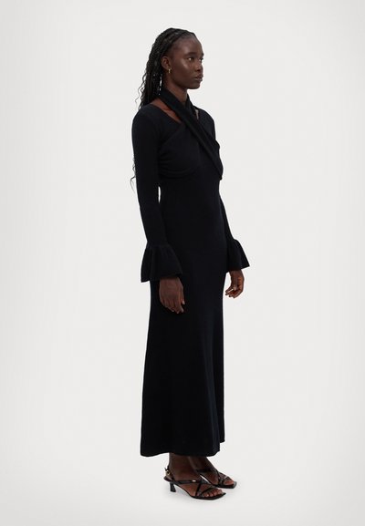 Long black dress with a fitted silhouette, draped cross-over neck, and flared sleeves. Worn with black strappy sandals. Smooth texture.